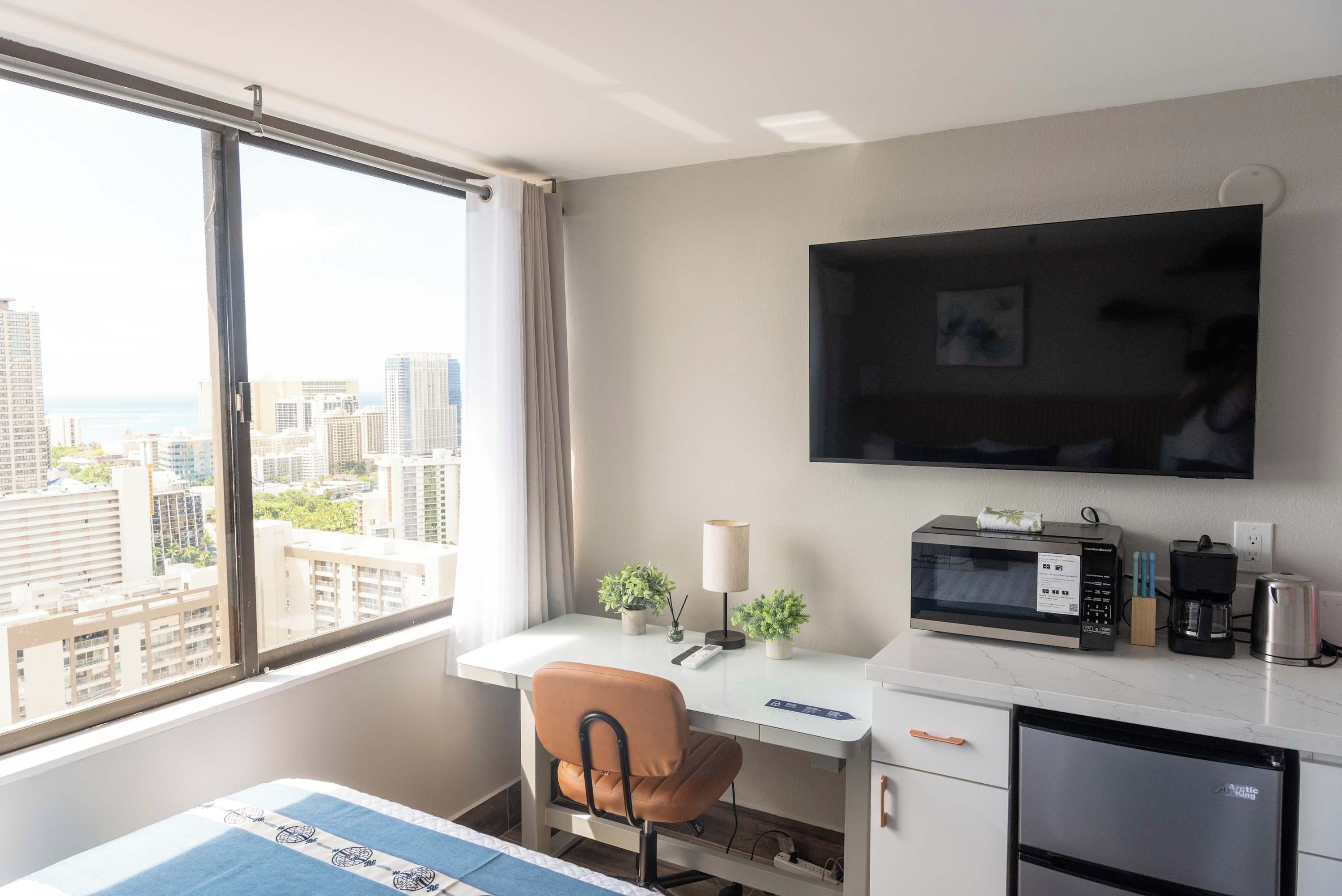 A hotel room with a desk, chair, bed, TV, microwave, coffee maker, and a view of the city.