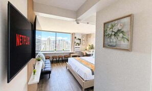 A modern hotel room with a large window, a bed, a desk, a chair, a sofa, and a TV screen displaying the Netflix logo. - Waikiki 2BR 2BA 34th Fl Ocean View Sunset/Sunrise (Honolulu)