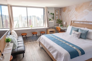A modern bedroom with a city view, a bed with a blue and white quilt, a desk with a lamp, and a seating area with a sofa and stools. - Waikiki 2BR 2BA 34th Fl Ocean View Sunset/Sunrise (Honolulu)