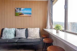 A modern living space with a black leather sofa, three cushions, a city view through a large window, and a small potted plant on the windowsill. - Waikiki 2BR 2BA 34th Fl Ocean View Sunset/Sunrise (Honolulu)