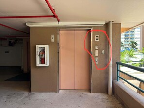 An elevator with a fire extinguisher mounted on the wall, a door with a number plate, and a view of a building with palm trees. - Waikiki 2BR 2BA 34th Fl Ocean View Sunset/Sunrise (Honolulu)