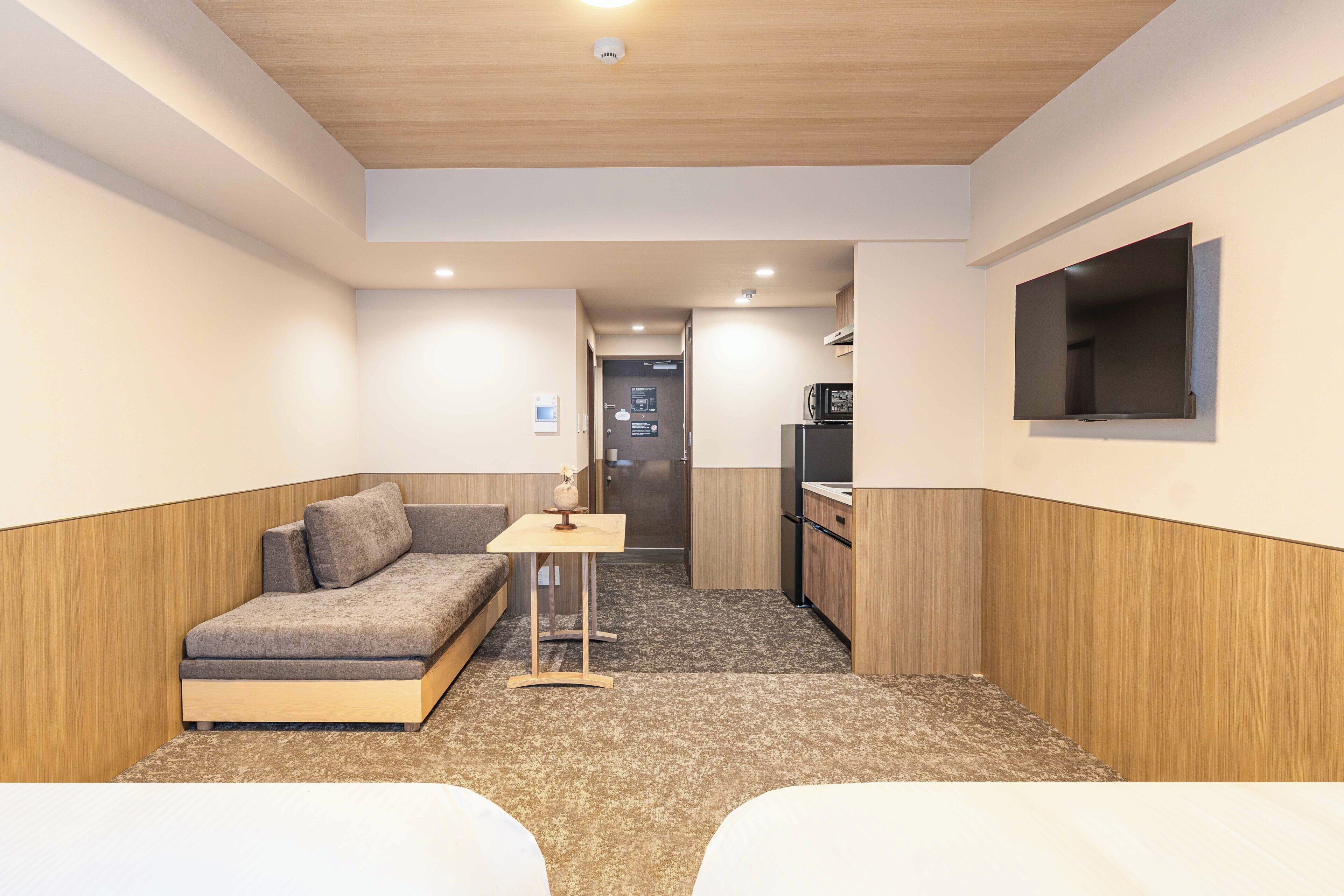 A compact hotel room with a sofa, a small table, a TV mounted on the wall, and a kitchenette.