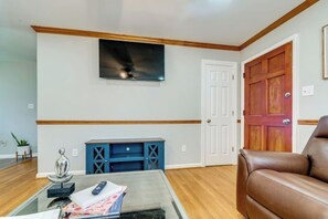 A living room with a television mounted on the wall, a wooden door, a brown leather sofa, and a blue cabinet with drawers. - Spacious 4BR/2.5BA Pet-Friendly Home in Clayton, NC – Minutes from Raleigh, Sleeps 8 (Clayton)