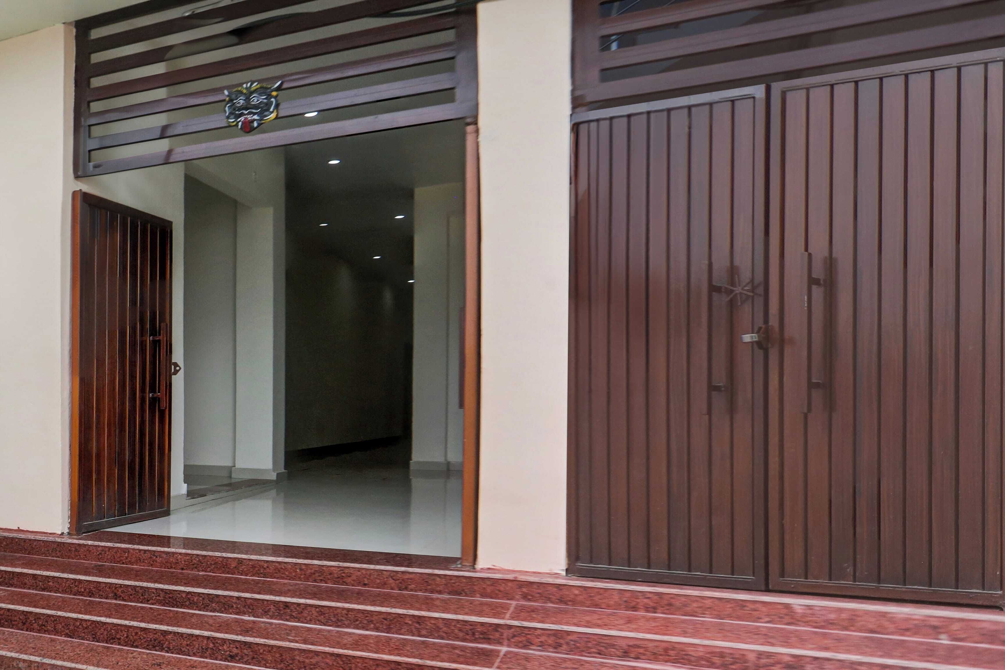 Entrada de um edifício com um conjunto de degraus que levam a uma porta, uma porta grande de cor castanha com puxador de metal, e um pequeno emblema decorativo acima da porta.