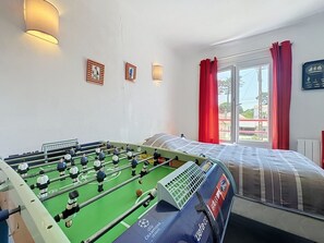 A room with a foosball table, a bed, a window with red curtains, and wall-mounted lights. - Claire Joie by Interhome (Saint Brevin les Pins)