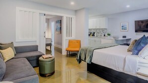 A modern hotel room with a grey sofa, a bed with white bedding, a small orange chair, and a kitchenette in the background. - The Montrose Hideaway, Downtown, Medical Center 07 (Houston)