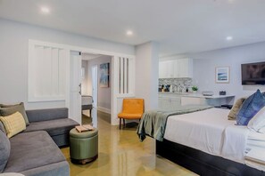 Ein modernes Hotelzimmer mit grauem Sofa, einem Bett mit weißer Bettwäsche, einem kleinen orangefarbenen Sessel und einer Küchenzeile im Hintergrund.