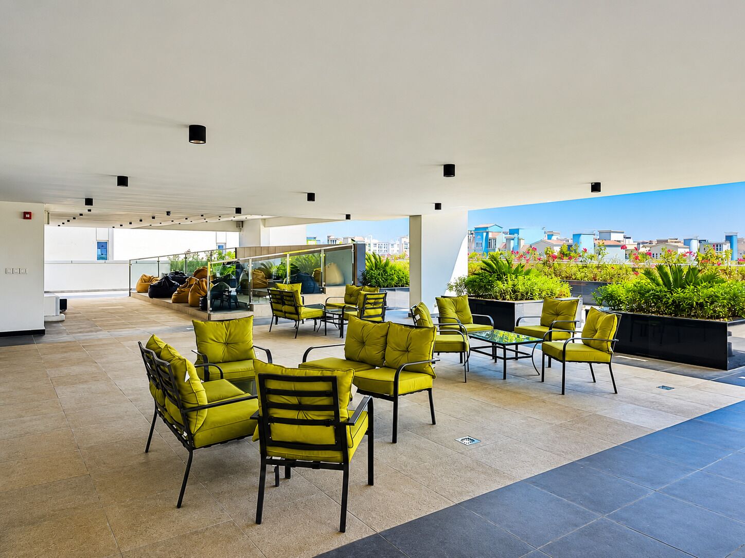 A rooftop terrace with yellow chairs and tables, a glass railing, and a view of buildings and greenery.