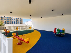 A children's play area with various play equipment, a white fence, and a building in the background. - Pearlz By Danube Residences - Al Furjan (Dubai)