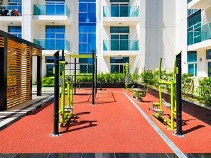 Ein Outdoor-Fitnessbereich mit Trainingsgeräten, umgeben von einem roten Gummiboden und neben einem Gebäude mit Balkonen und großen Fenstern.