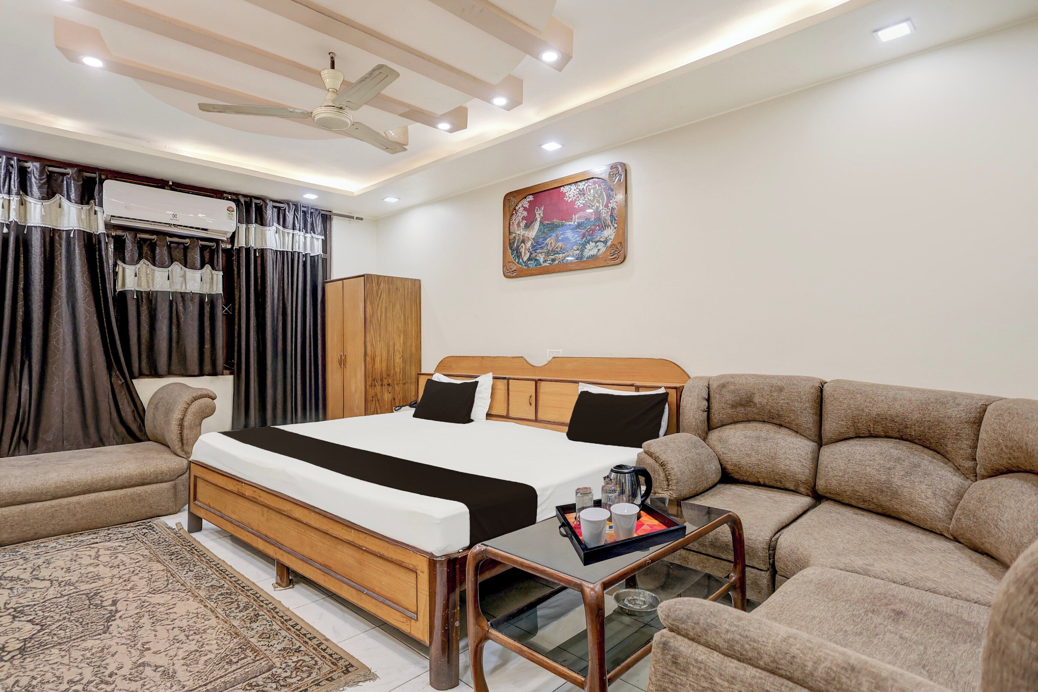 A hotel room with a bed, a sofa, a coffee table, a painting on the wall, and a ceiling fan.
