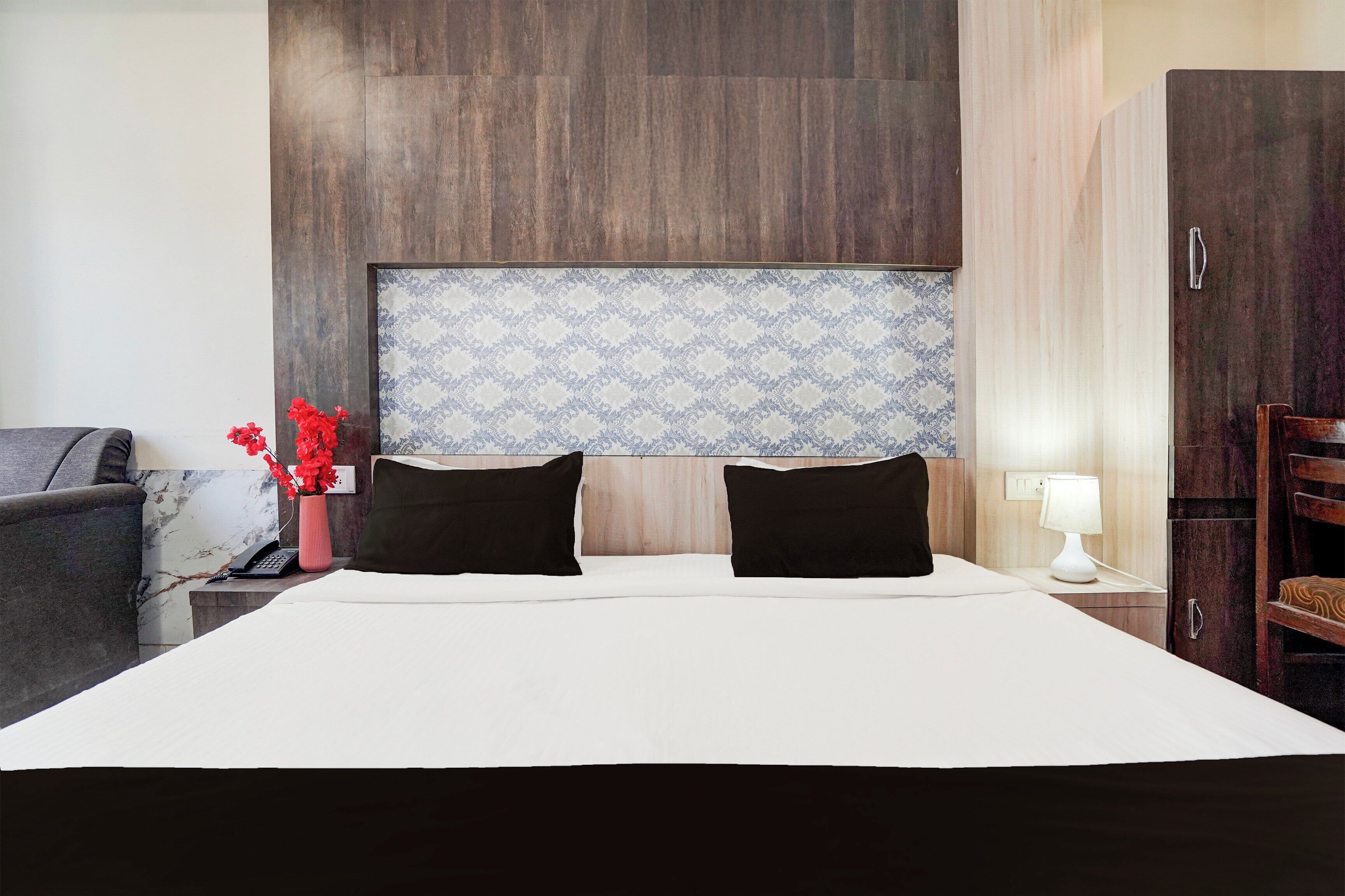 Hotelkamer met een groot bed, twee zwarte kussens, een nachtkastje met een lamp, een vaas met rode bloemen en een houten garderobe.