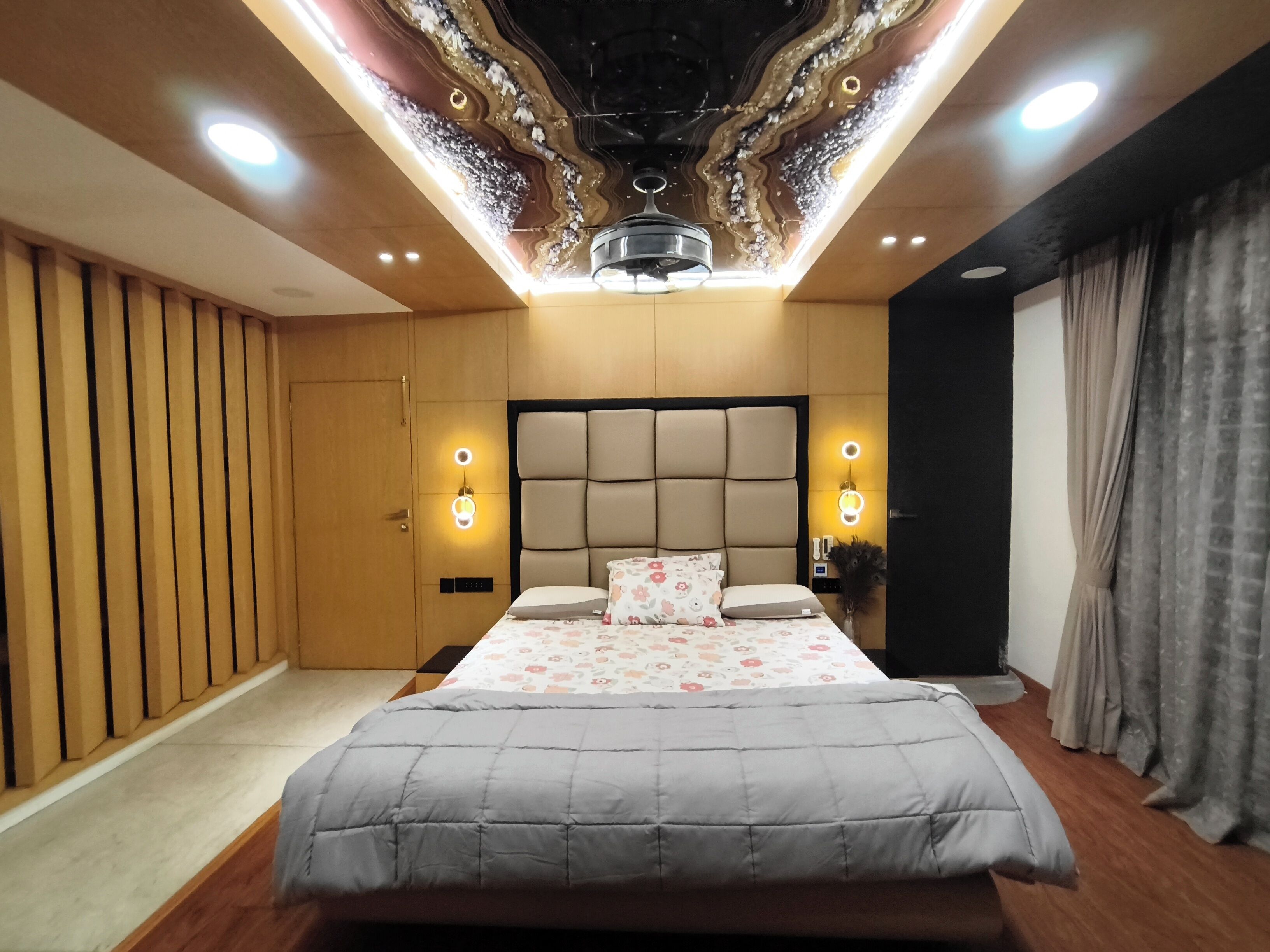 A modern bedroom with a tufted headboard, a large bed with a grey quilt, two wall-mounted reading lights, and a ceiling with a decorative light fixture.