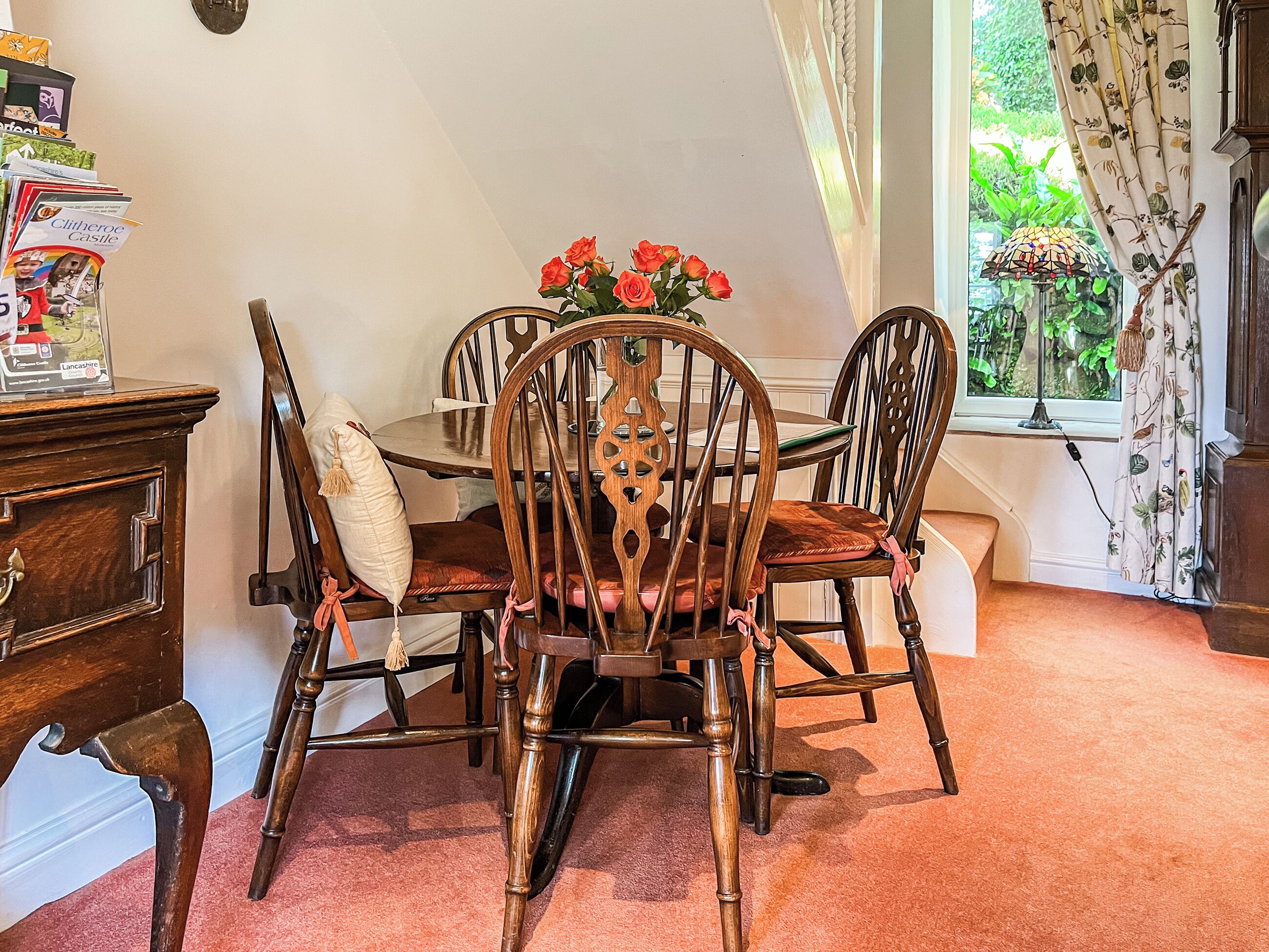 Un comedor con mesa y sillas de madera, un jarrón con flores y una ventana con cortinas.