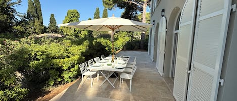 Un patio al aire libre con una mesa lista para comer, rodeado de follaje y un edificio con persianas blancas.