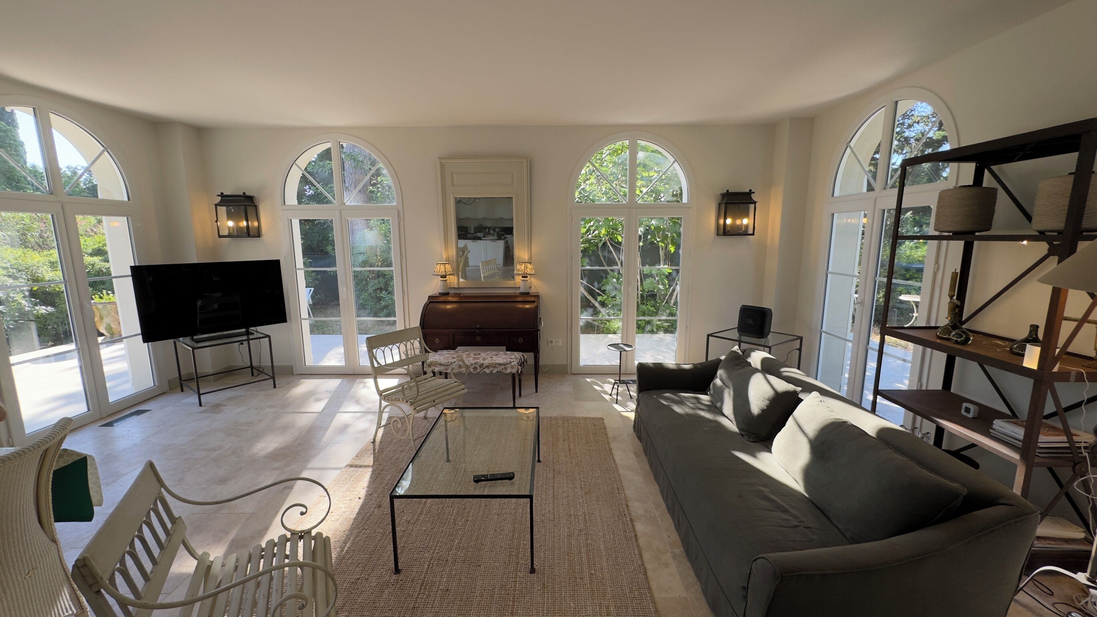 A spacious living room with a sofa, a coffee table, and a TV. There are large windows and a view of greenery outside.