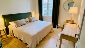 Et hotelværelse med seng, skrivebord, stol, lampe, et vindue med gardiner og et spejl på væggen.