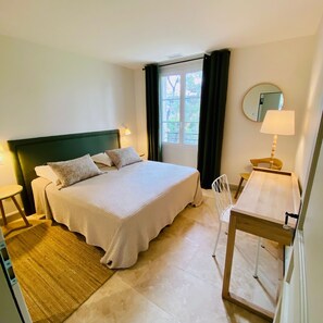 Ein Hotelzimmer mit Bett, Schreibtisch, Stuhl, Lampe, einem Fenster mit Vorhängen und einem Spiegel an der Wand.