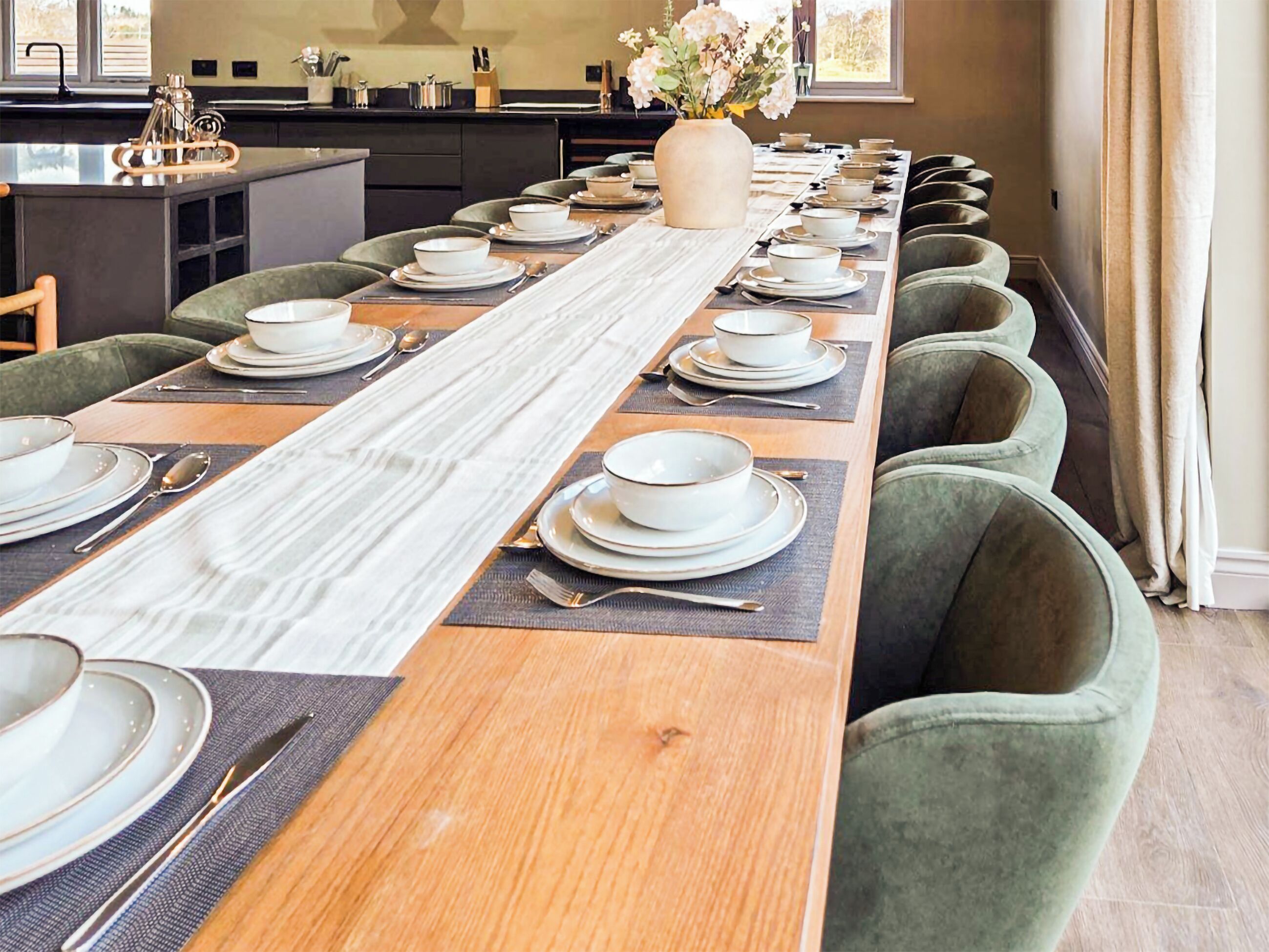 A long dining table set with plates, bowls, and cutlery, flanked by green upholstered chairs.