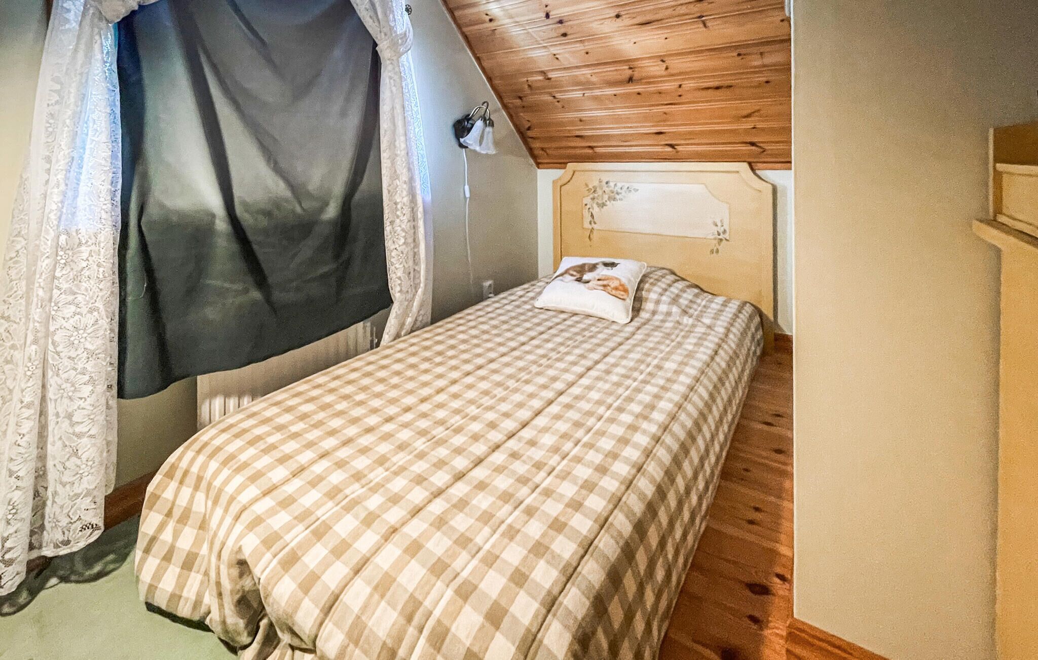 A bedroom with a checkered bedspread, a pillow with a cat design, and a wooden ceiling.