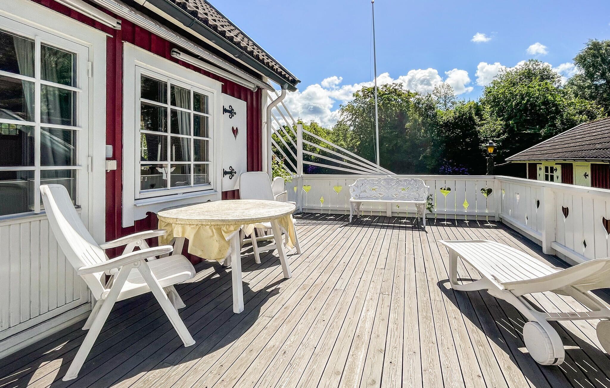 Une terrasse en bois avec une table ronde et des chaises blanches, un bâtiment blanc orné de bordures rouges et un ciel bleu dégagé.