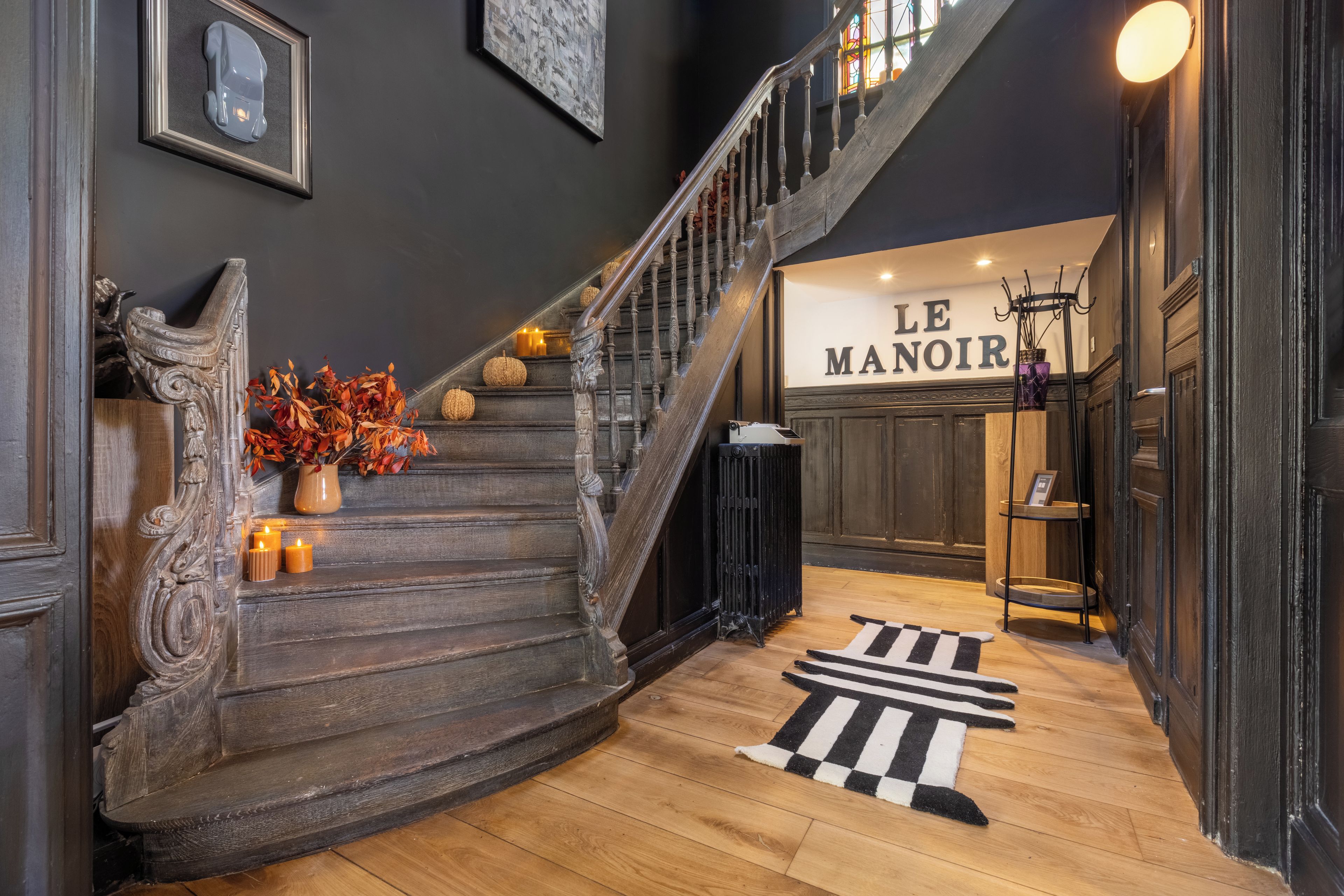 Un grand escalier au revêtement en bois foncé, un tapis rayé noir et blanc au pied de l’escalier et, en arrière-plan, une enseigne indiquant « LE MANOIR ».