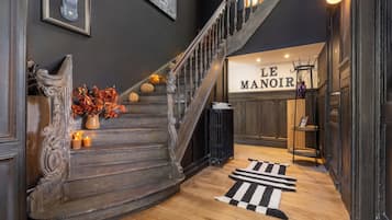 Een statige trap met een donkerhouten afwerking, een zwart-wit gestreept tapijt aan de voet ervan, en een bord met de tekst 'LE MANOIR' op de achtergrond.