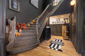 A grand staircase with a dark wooden finish, a black and white striped rug at the bottom, and a sign reading 'LE MANOIR' in the background.