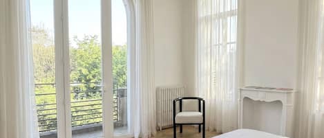 A bedroom with a large sliding glass door leading to a balcony, a chair, a radiator, and a fireplace mantel.