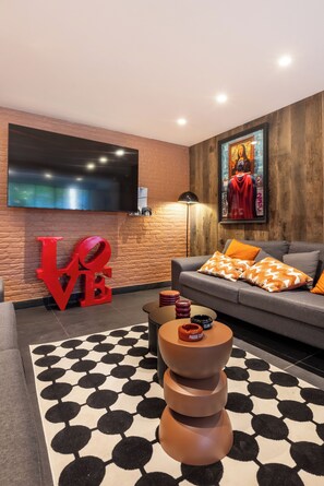 A modern living room with a flat-screen TV, a large red 'LOVE' sign, a sofa with patterned cushions, and a unique coffee table.
