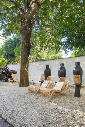 A patio area with wicker lounge chairs, a tree, and decorative vases against a white brick wall.