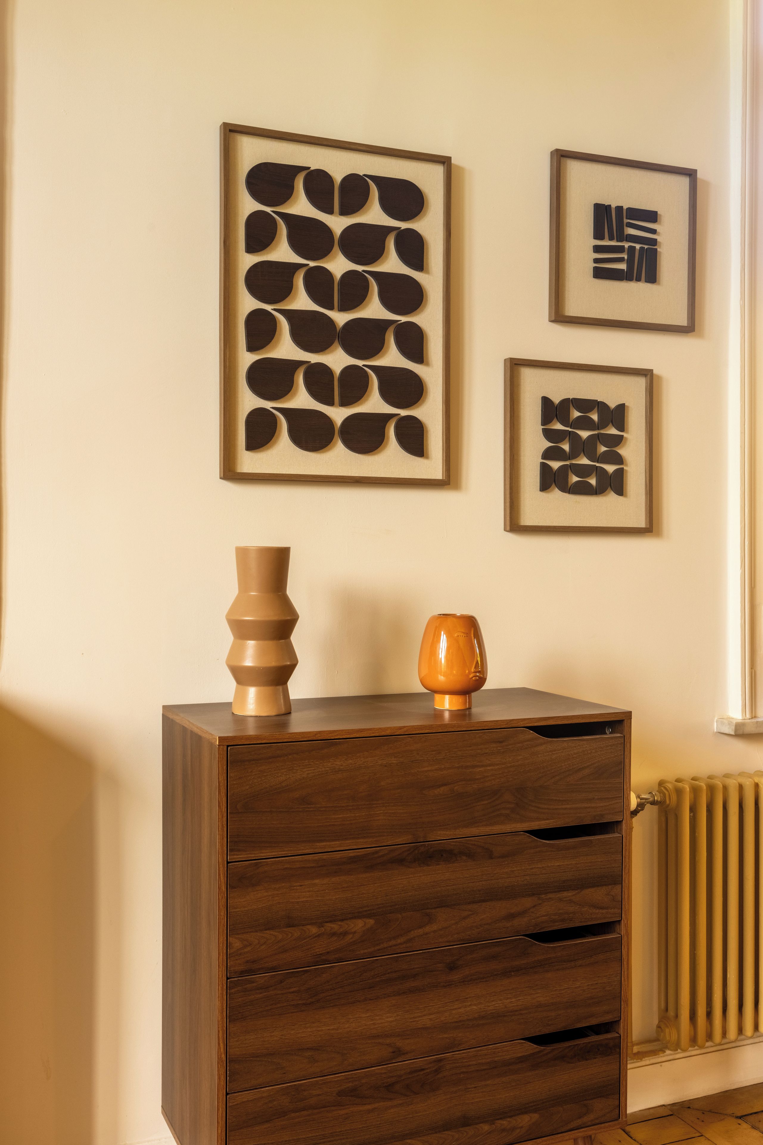 Un meuble en bois avec des tiroirs, un vase et une œuvre d’art abstraite accrochée au mur.