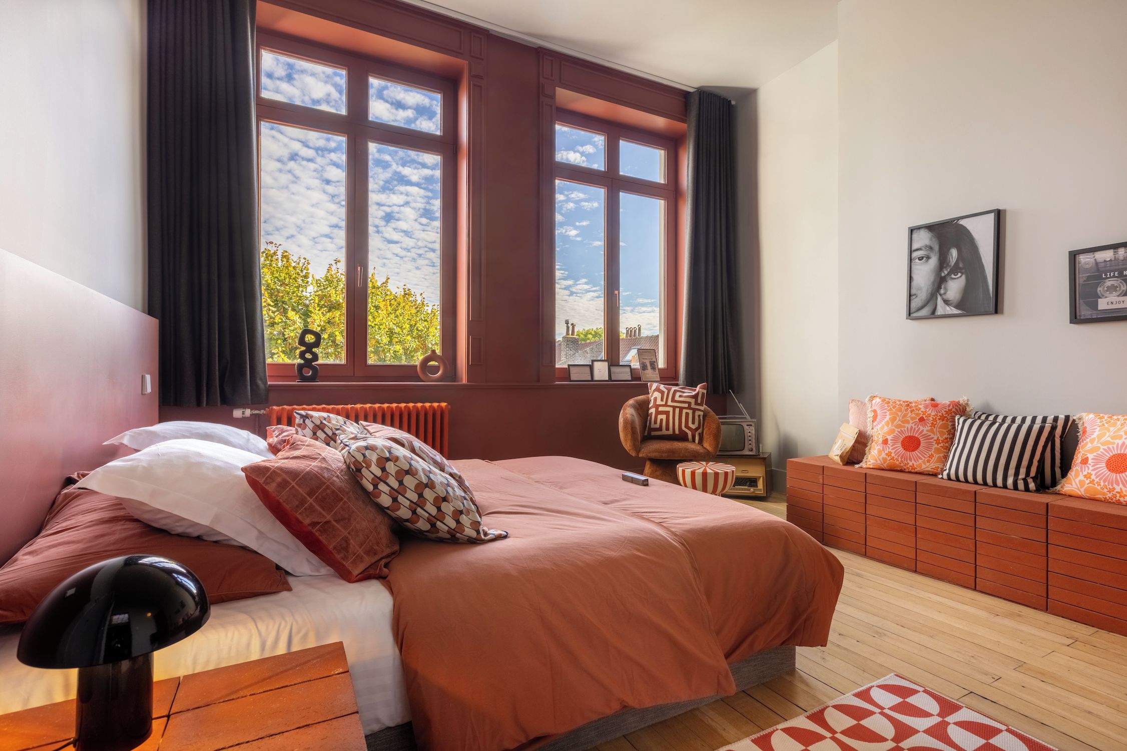A bedroom with a large bed, a chair, a sofa, and a window with a view of trees and buildings.