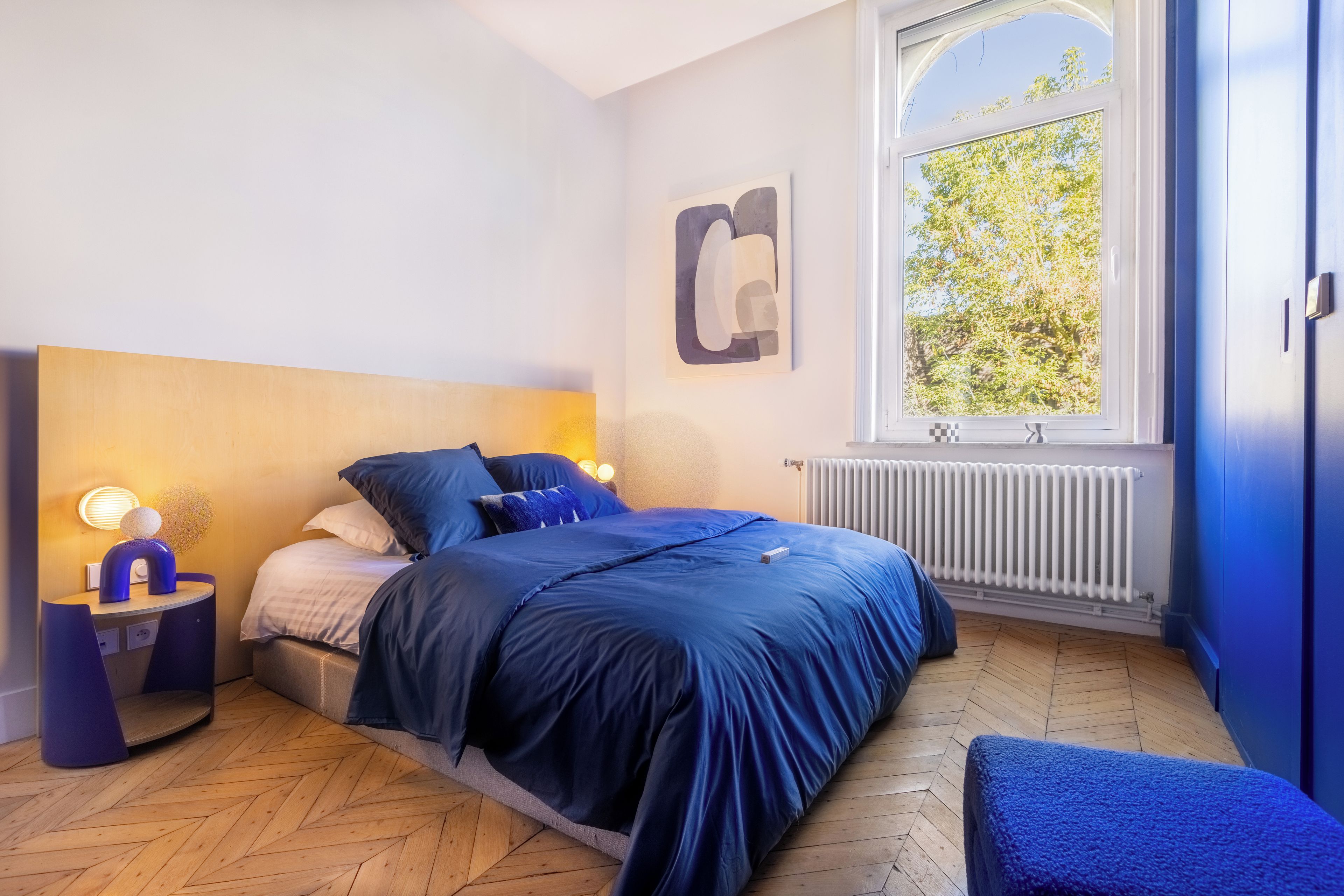 Une chambre à coucher avec un lit, une table de chevet, une lampe et une fenêtre donnant sur des arbres.
