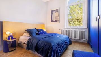 Une chambre à coucher avec un lit, une table de chevet, une lampe et une fenêtre donnant sur des arbres.