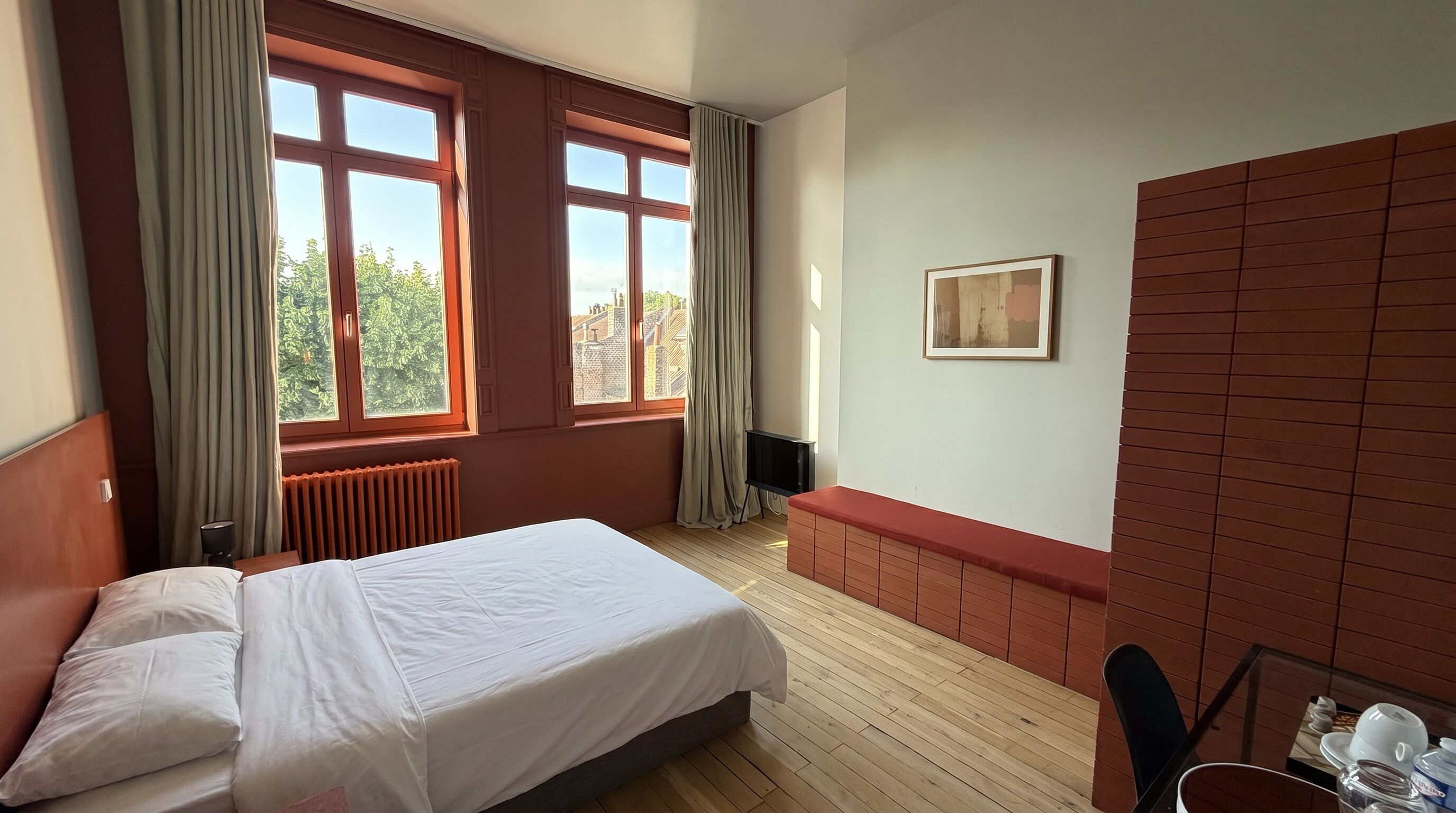 Een hotelkamer met een bed, een radiator, een raam met gordijnen, een televisie en een schilderij aan de muur.