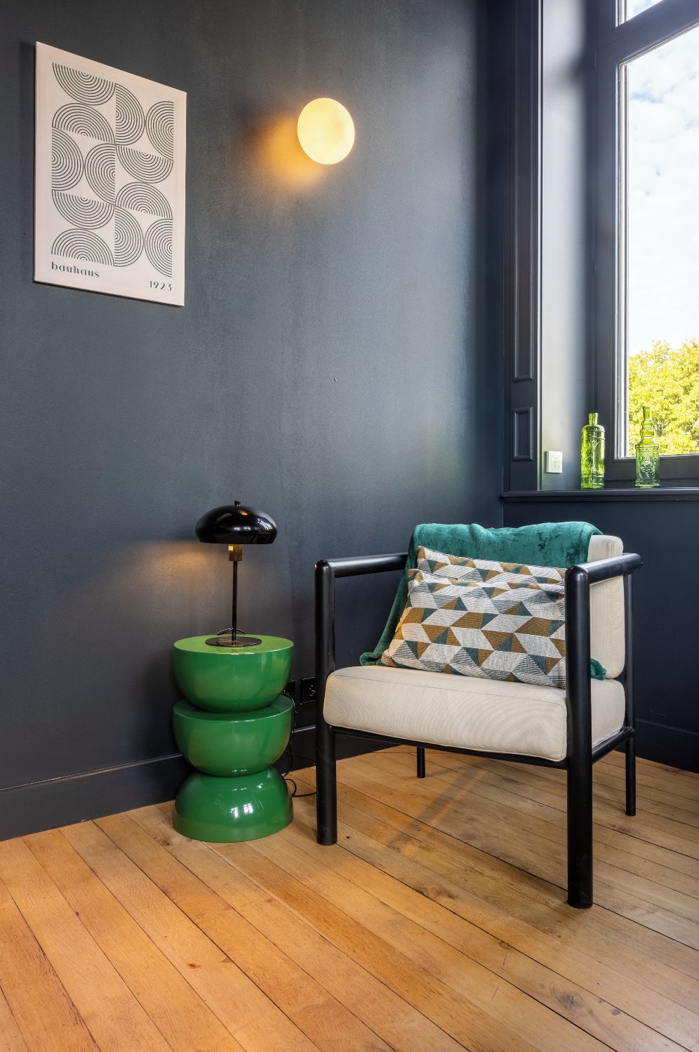 Une pièce avec un mur bleu foncé, un lampadaire vert, une chaise avec un coussin à motifs et une fenêtre donnant sur des arbres.