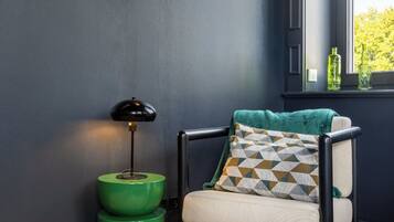 Une pièce avec un mur bleu foncé, un lampadaire vert, une chaise avec un coussin à motifs et une fenêtre donnant sur des arbres.