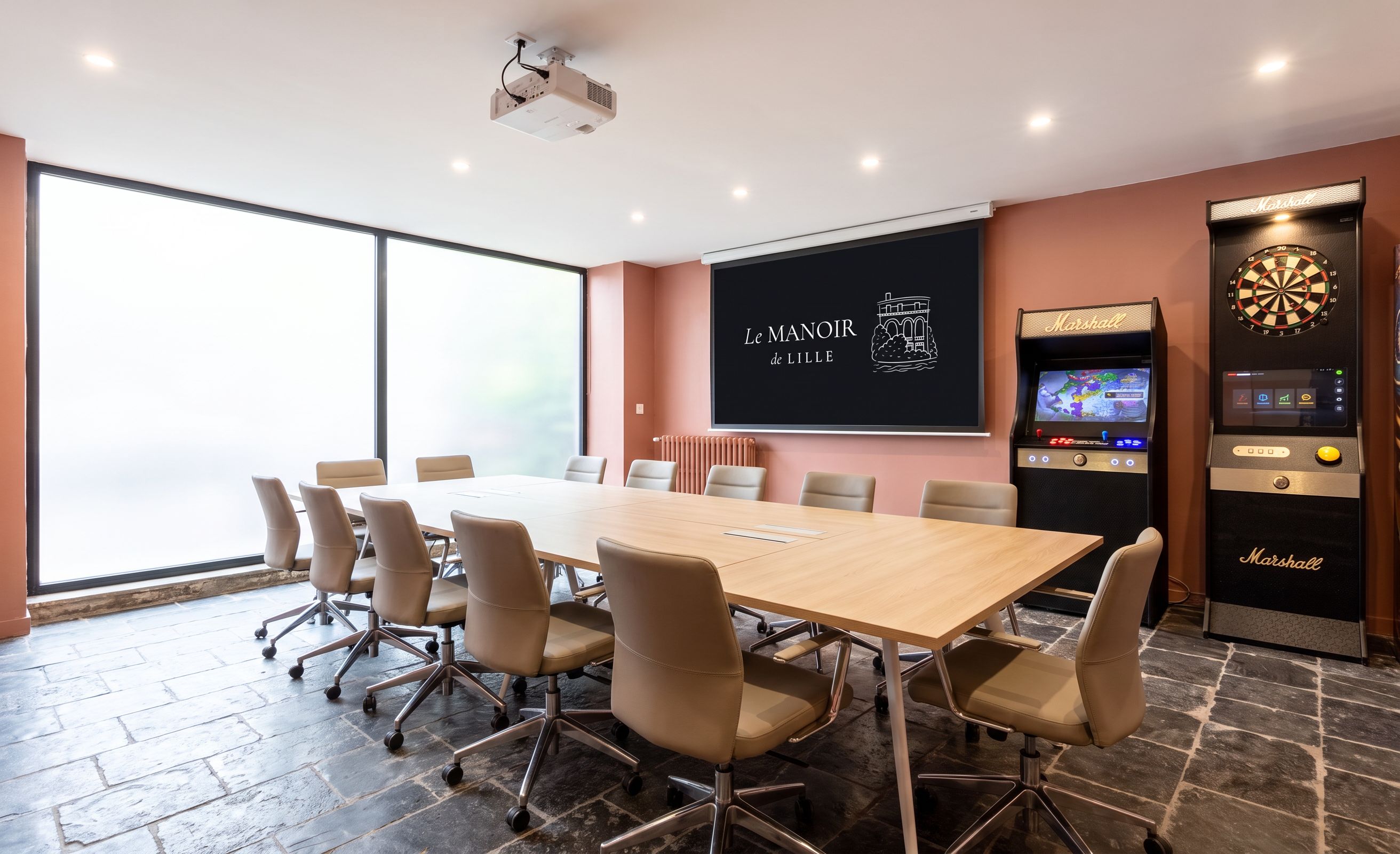 Une salle de conférence avec une longue table, des chaises, un projecteur, un jeu de fléchettes et un écran affichant le texte « Le Manoir de Lille ».