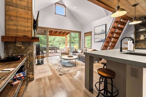 A spacious kitchen with a stone fireplace, wooden beams, and a large window overlooking a deck.