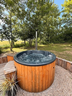 Een hottub omgeven door een houten terras en grind, met bomen op de achtergrond.