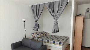 Un dormitorio pequeño con cama, cortinas grises, un sofá gris, una lámpara de pie y un armario de madera.