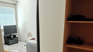 Una habitación con piso de madera, pared blanca, televisor sobre un mueble y estante con dispositivos electrónicos.