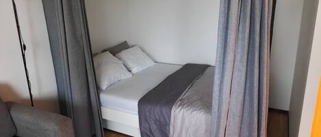 A bedroom with a bed, grey curtains, a bedside lamp, and a wooden floor.