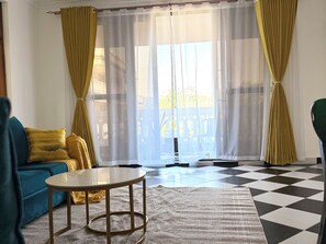 A room with a checkered floor, a blue sofa, a round table, and yellow curtains. - 2-bedroom apartment with AC, 5 mins from Diani Beach (Diani Beach)