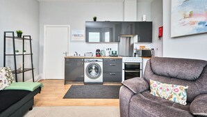 A compact living space with a kitchenette, a washing machine, a sofa, and a painting on the wall. - No.1 West Beach Apartments - UK52002 (Portrush)