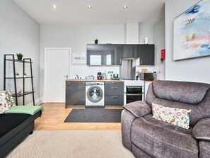 A compact living space with a kitchenette, a washing machine, a sofa, and a painting on the wall. - No.1 West Beach Apartments - UK52002 (Portrush)