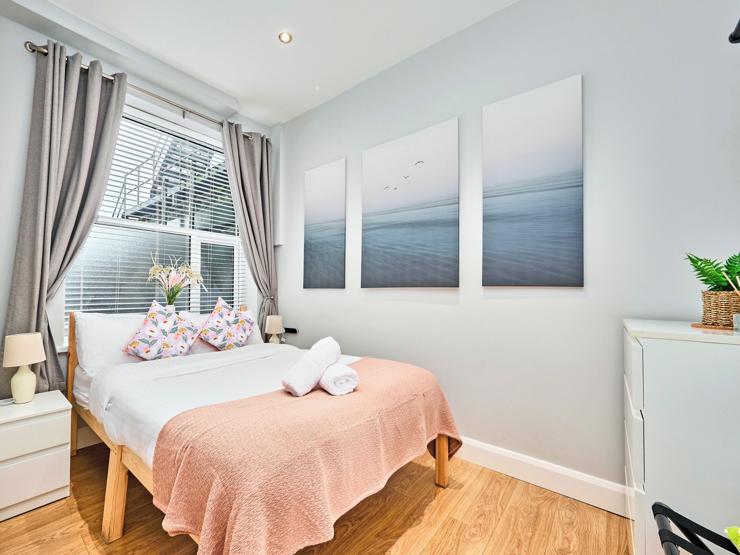 Ein Schlafzimmer mit Bett, Nachttisch, Lampe, einem Fenster mit Vorhängen und Wandbildern.