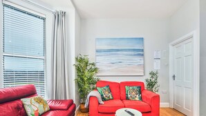 A red sofa with tiger-themed cushions, a small white table with a remote, and a painting of a beach scene. - No.3 West Beach Apartments - UK52003 (Portrush)
