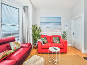 A red sofa with tiger-themed cushions, a small white table with a remote, and a painting of a beach scene. - No.3 West Beach Apartments - UK52003 (Portrush)